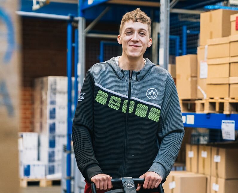 Ein Mann schiebt einen Wagen. Er ist in einer Lagerhalle.